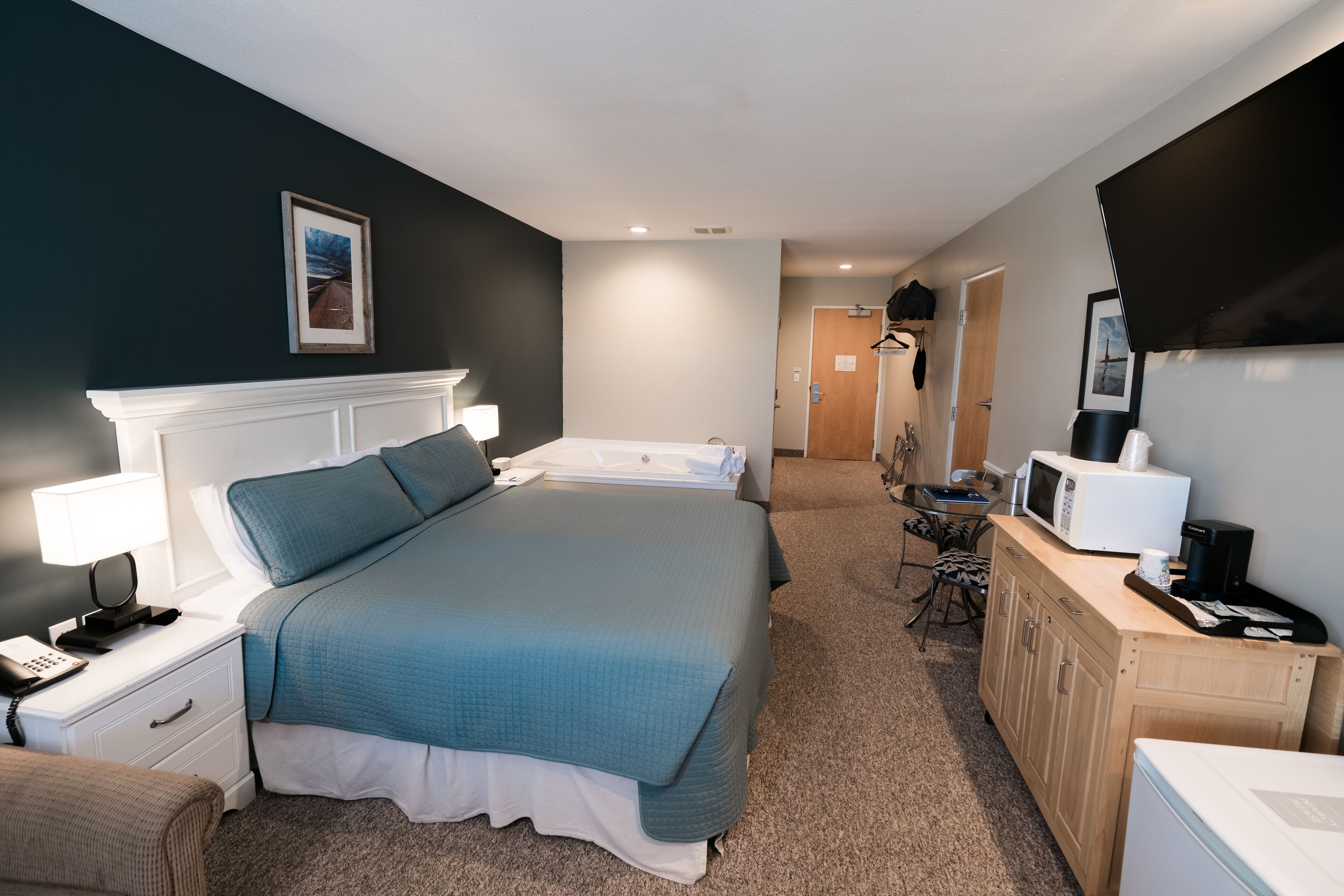 Habitación de hotel con una cama grande, mesita de noche con lámpara, microondas y una pequeña zona de kitchenette.