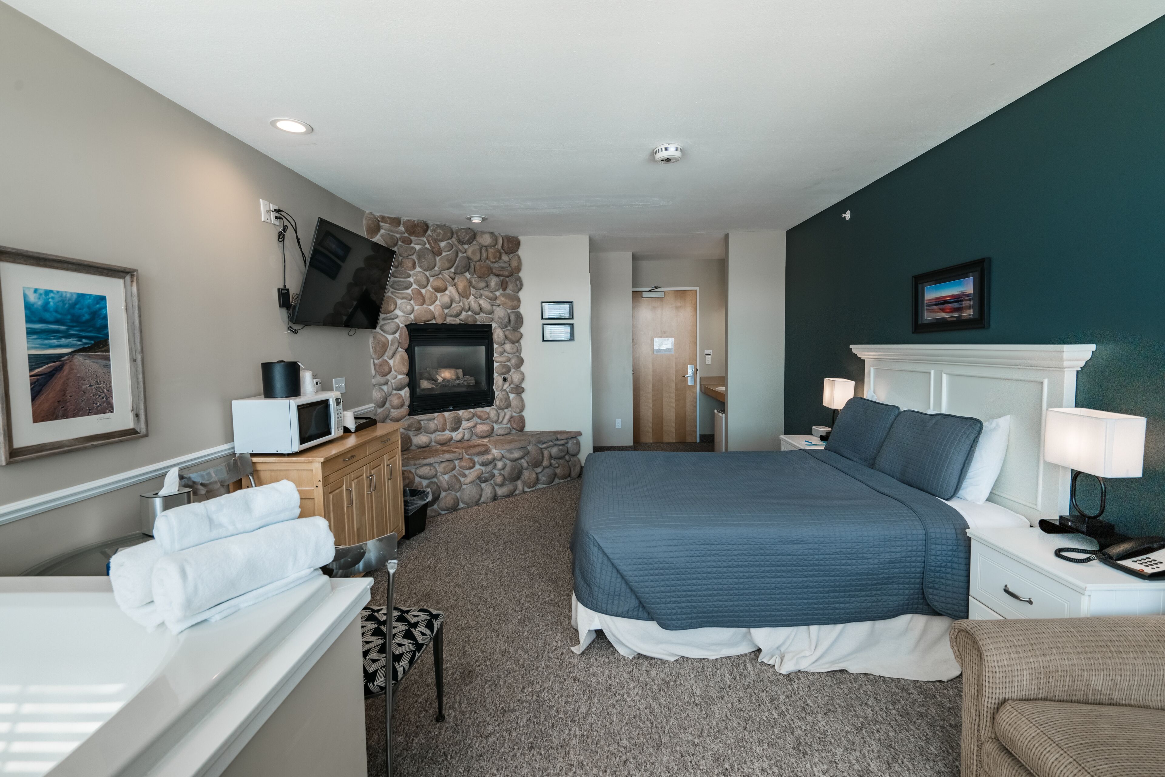 A hotel room with a stone accent wall, a bed with a blue quilt, a white bedside table with a lamp, a microwave, and a TV mounted on the wall.