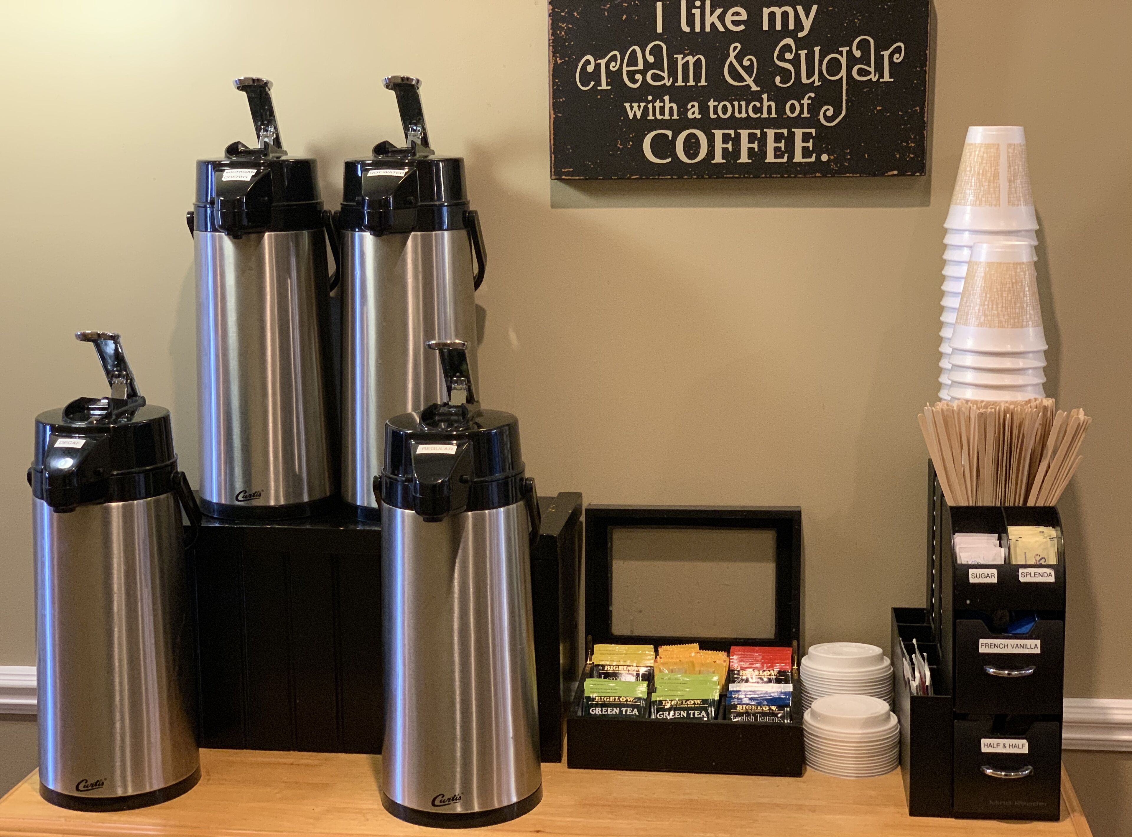 A coffee station with multiple dispensers, a sign, and a sugar and cream dispenser.