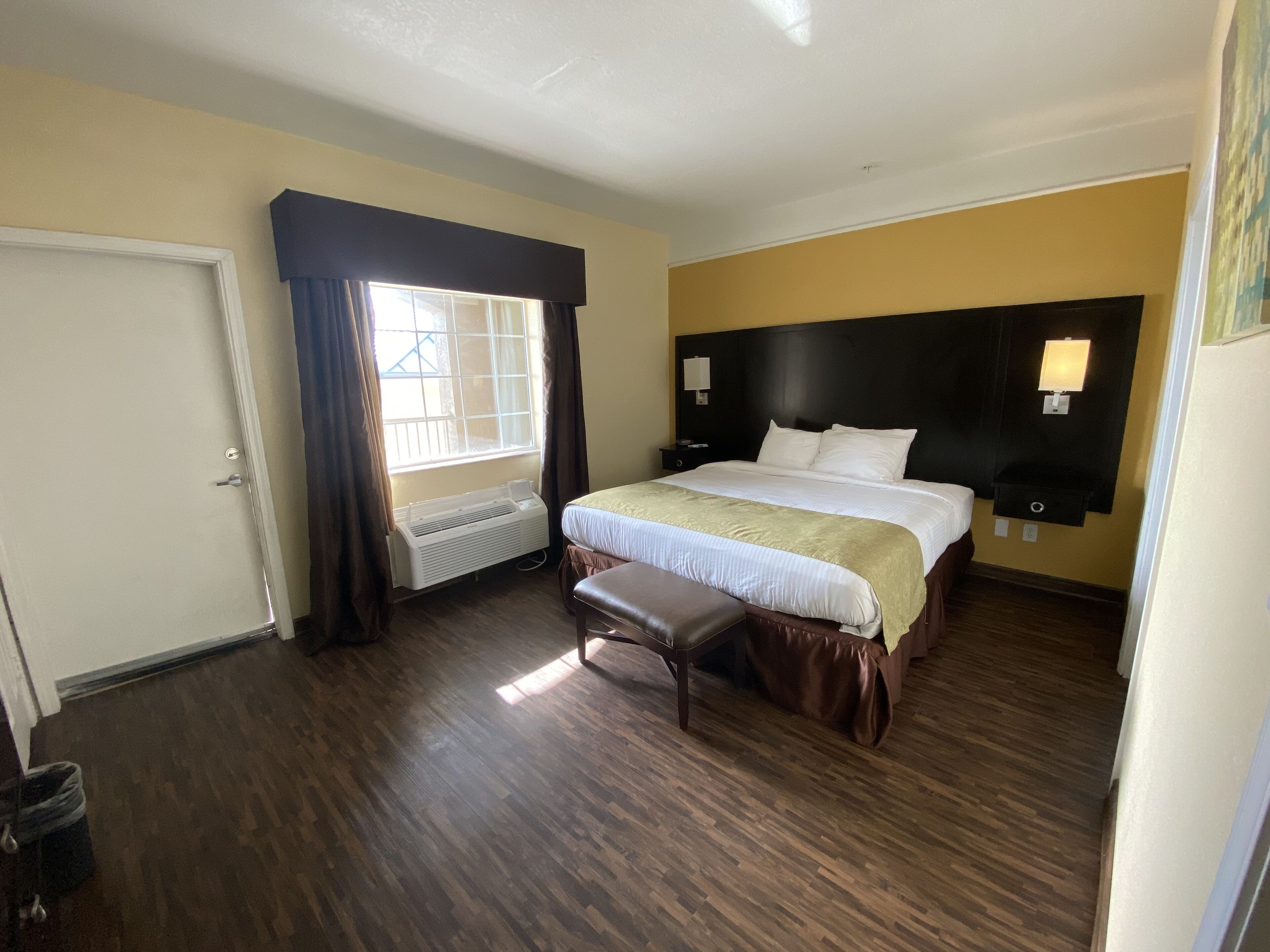 A hotel room with a large bed, a bedside table, a nightstand, a window with curtains, a wall-mounted lamp, and a radiator.