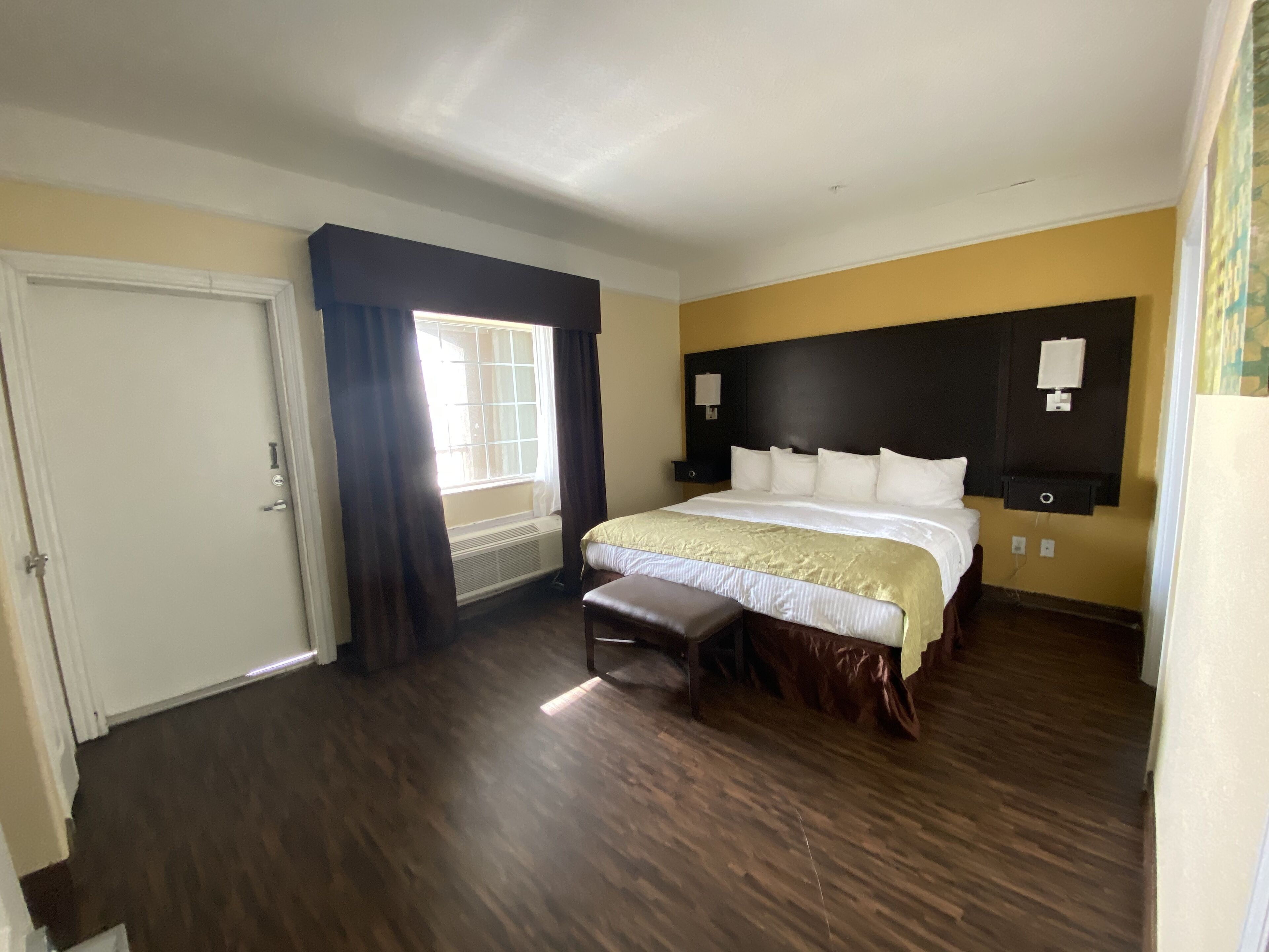 Ein Hotelzimmer mit einem großen Bett, einer Sitzbank, einem fenster mit Vorhängen, einer Wandheizung und einer Tür.
