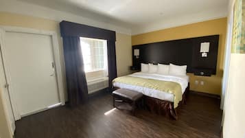 Une chambre d’hôtel avec un grand lit, un banc, une fenêtre avec des rideaux, un radiateur fixé au mur et une porte.