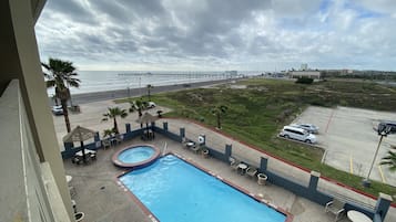 A swimming pool with lounge chairs and a hot tub, surrounded by a parking lot and a view of the ocean and a bridge.