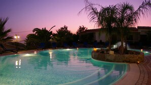 A swimming pool with a tiled edge, surrounded by lounge chairs and palm trees, under a twilight sky. - Hotel Village Eden (Ricadi)