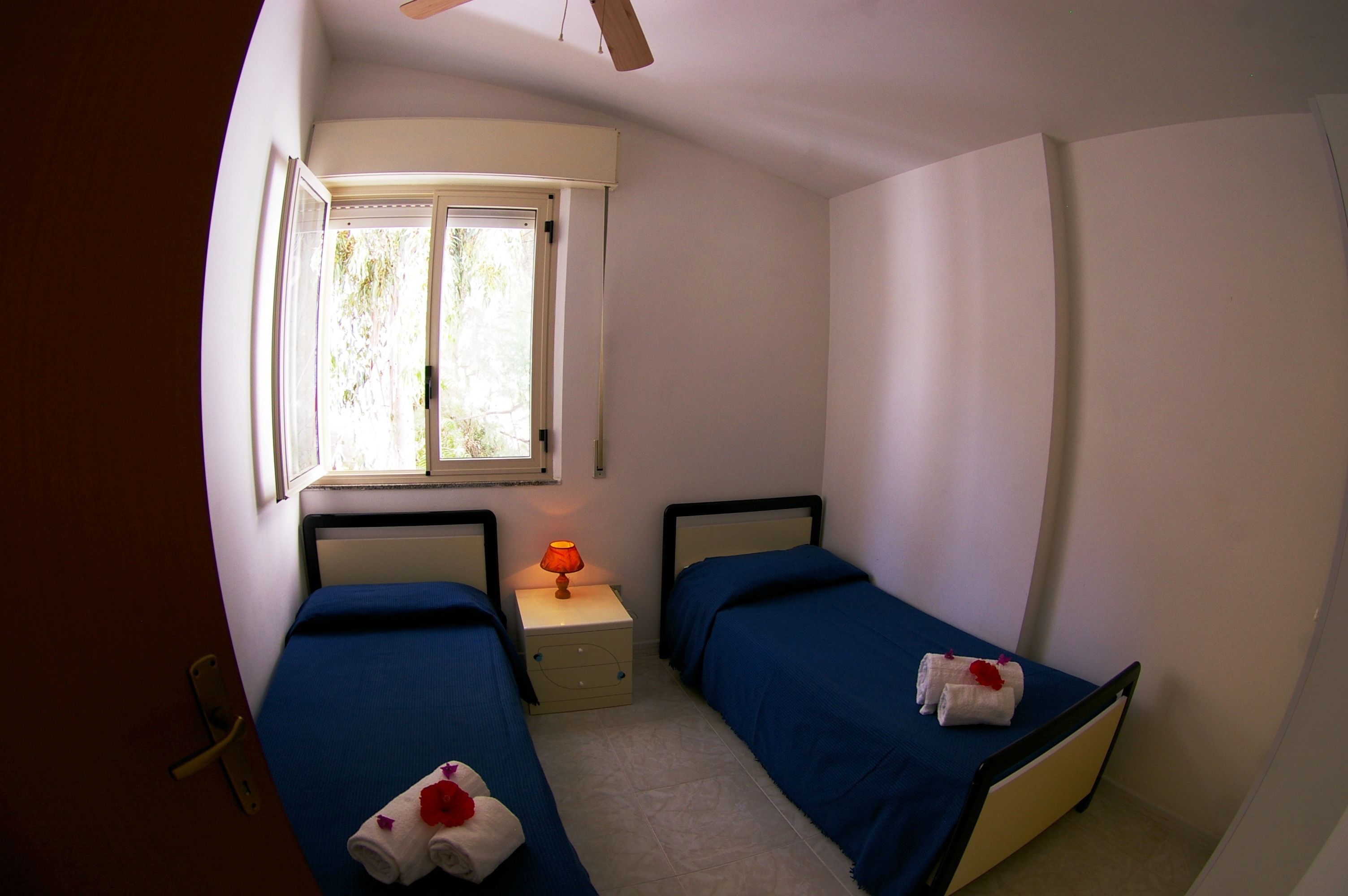 Une petite chambre bien éclairée, avec deux lits simples, une table de chevet avec une lampe et une fenêtre laissant entrer la lumière naturelle.