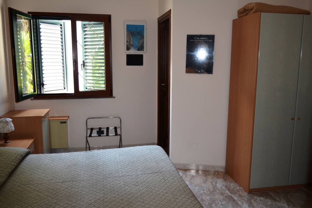 A bedroom with a bed, a wardrobe, a desk, a chair, and a window with blinds.