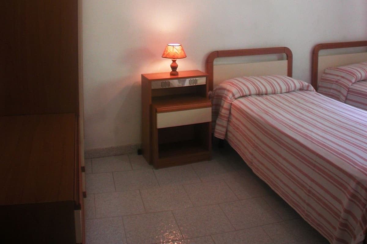 A bedroom with a wooden wardrobe, a bedside table with a lamp, two beds with striped bedding, and a ceiling fan.