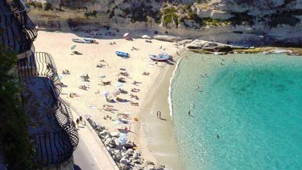 A coastal beach with a historic building on a cliff, clear turquoise waters, and a rocky shoreline. - Hotel Village Eden (Ricadi)