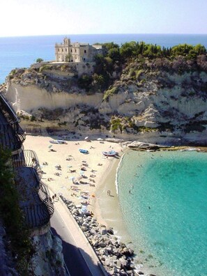 A coastal beach with a building on a cliff, clear turquoise waters, and rocky terrain. - Hotel Village Eden (Ricadi)
