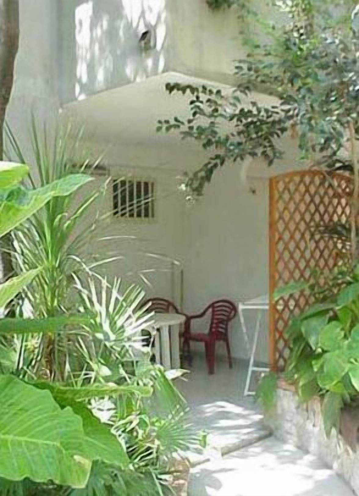 A small patio area with a table and two chairs, surrounded by lush green plants.