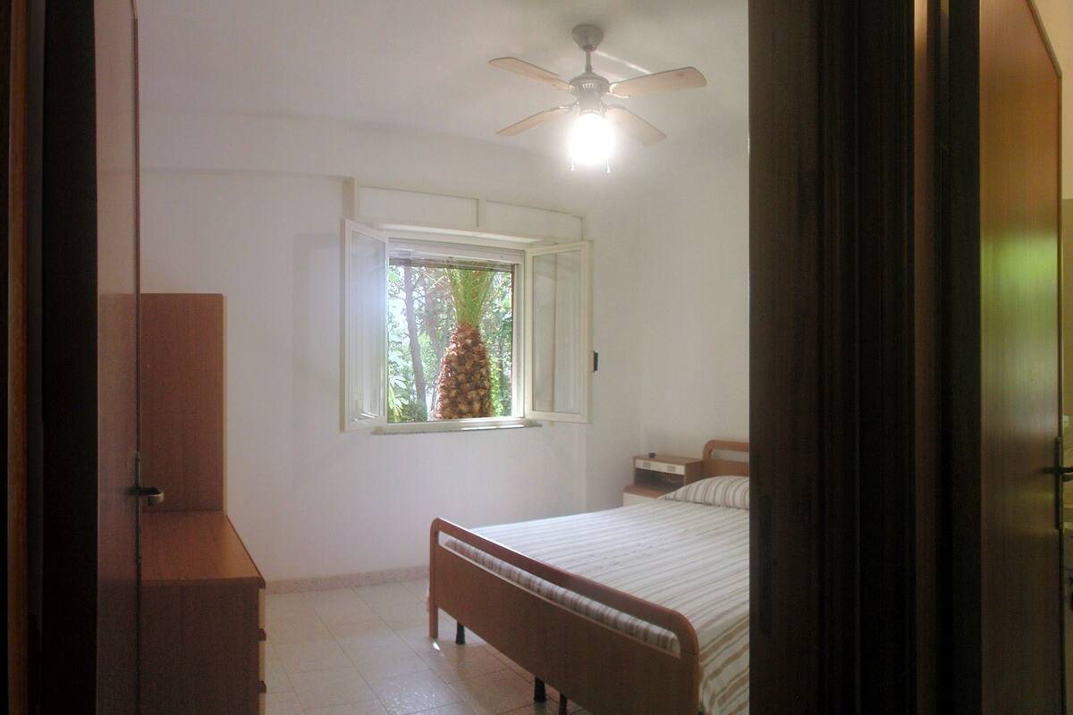 A bedroom with a bed, a wooden dresser, a window with a view of greenery, and a ceiling fan.
