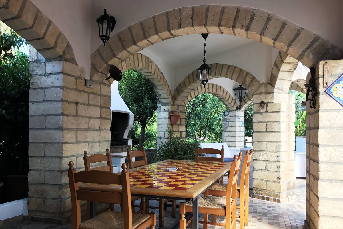 An outdoor patio with a checkered table and wooden chairs, surrounded by stone arches and greenery.