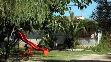 A playground with a red slide, surrounded by trees and a house in the background.