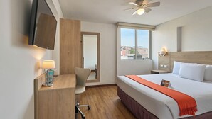 Habitación de hotel con cama, escritorio, silla, televisor, ventilador de techo, ventana y mesita de noche.