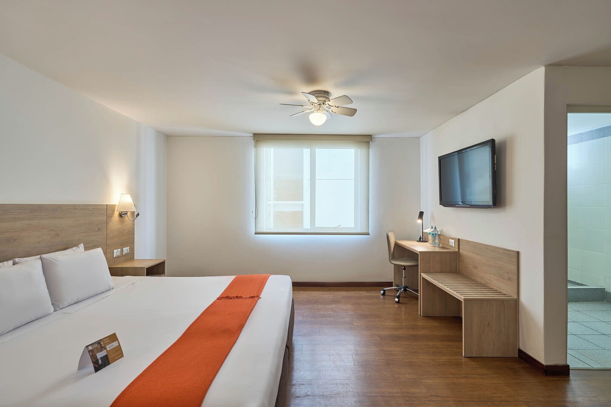 A hotel room with a bed, a desk, a chair, a TV, a ceiling fan, and a window.