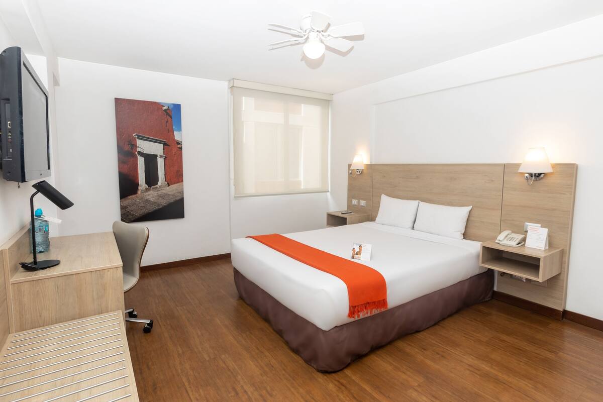 Ein Hotelzimmer mit Bett, Schreibtisch, Stuhl und einem an der Wand befestigten Fernseher.