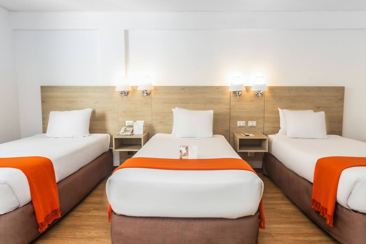 Ein Hotelzimmer mit drei Einzelbetten, jedes mit einem hölzernen Kopfteil und weißer Bettwäsche. Auf der linken Seite befinden sich zwei Nachttische mit Lampen und einem Telefon.