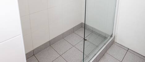 Un box de ducha con paredes de vidrio, suelo de baldosas blancas, barra de apoyo y perchero.
