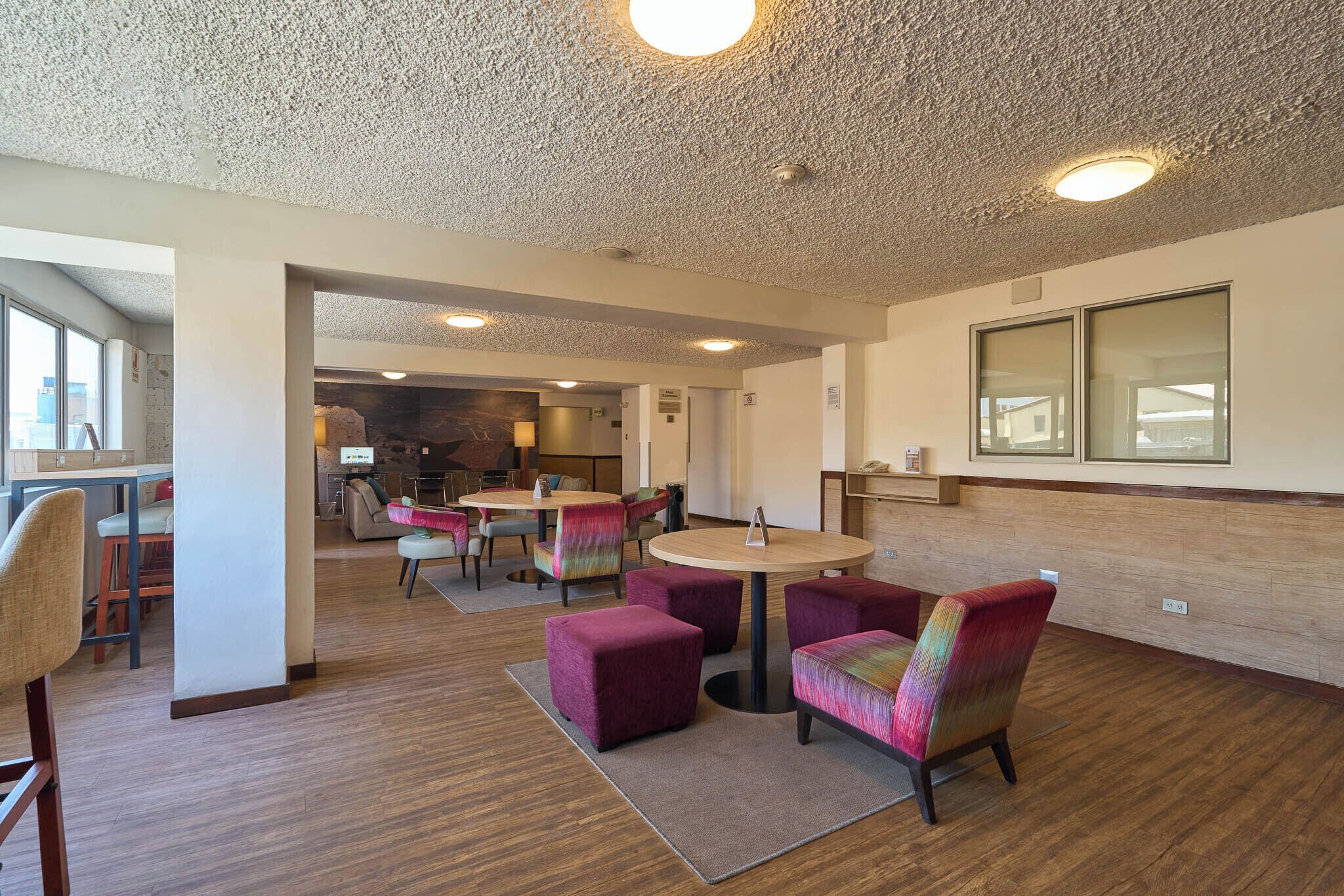A modern hotel lobby with round tables, cushioned chairs, a kitchen area, and a large window.
