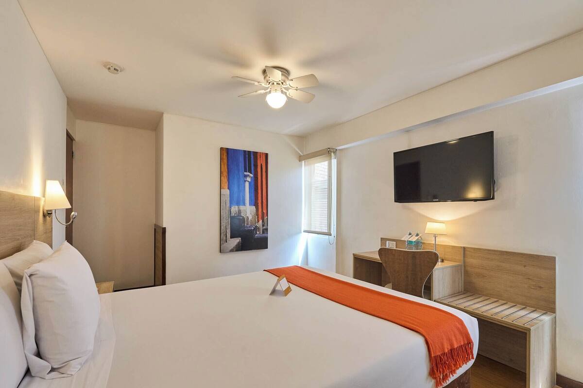 Ein Hotelzimmer mit Bett, Schreibtisch, Stuhl, Fernseher und Deckenventilator.