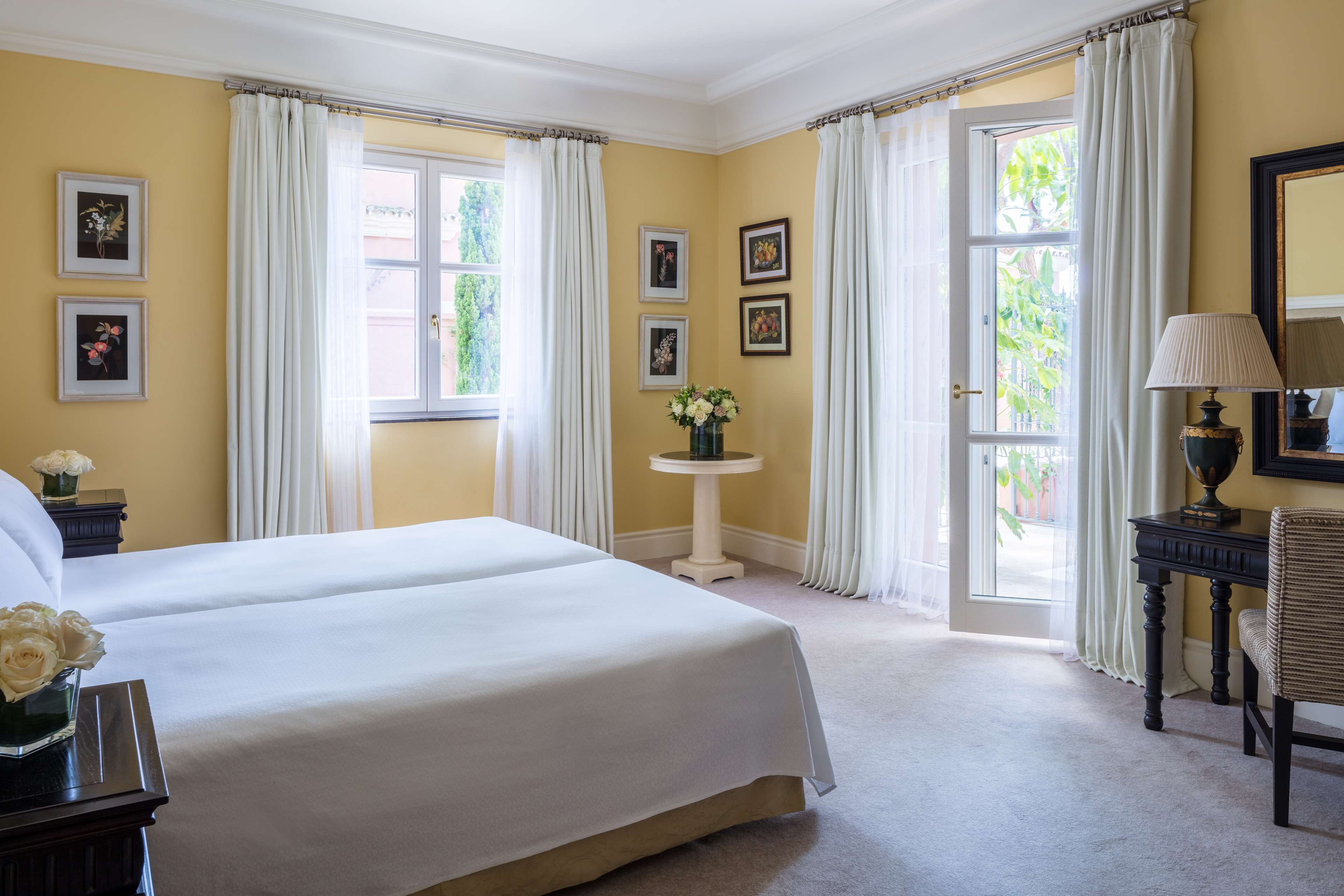 Quarto de hotel com duas camas, uma mesa redonda pequena com um vaso de flores, um abajur e quadros nas paredes.