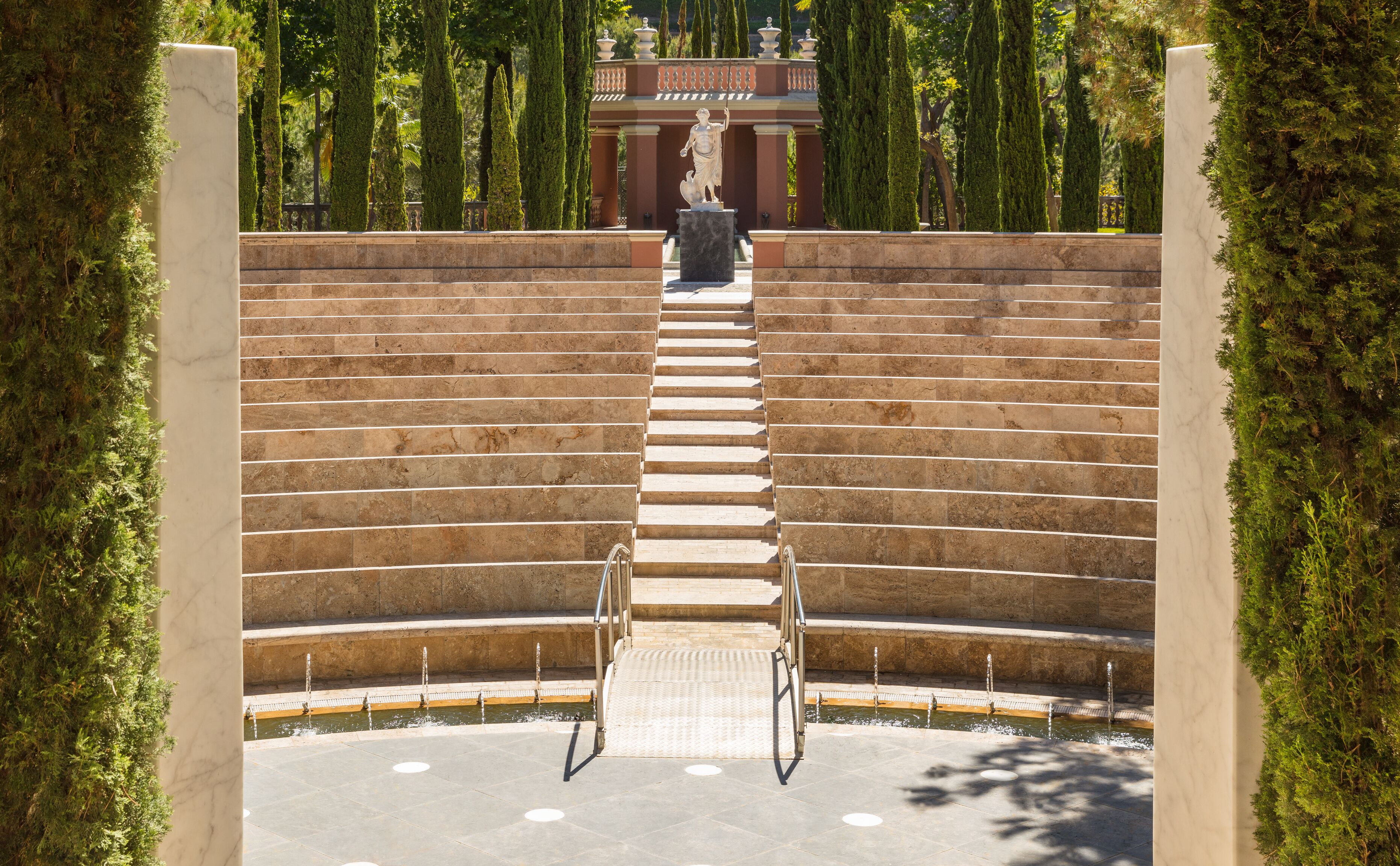 Una imponente escalera conduce a una estatua, rodeada de exuberante vegetación y una fuente.