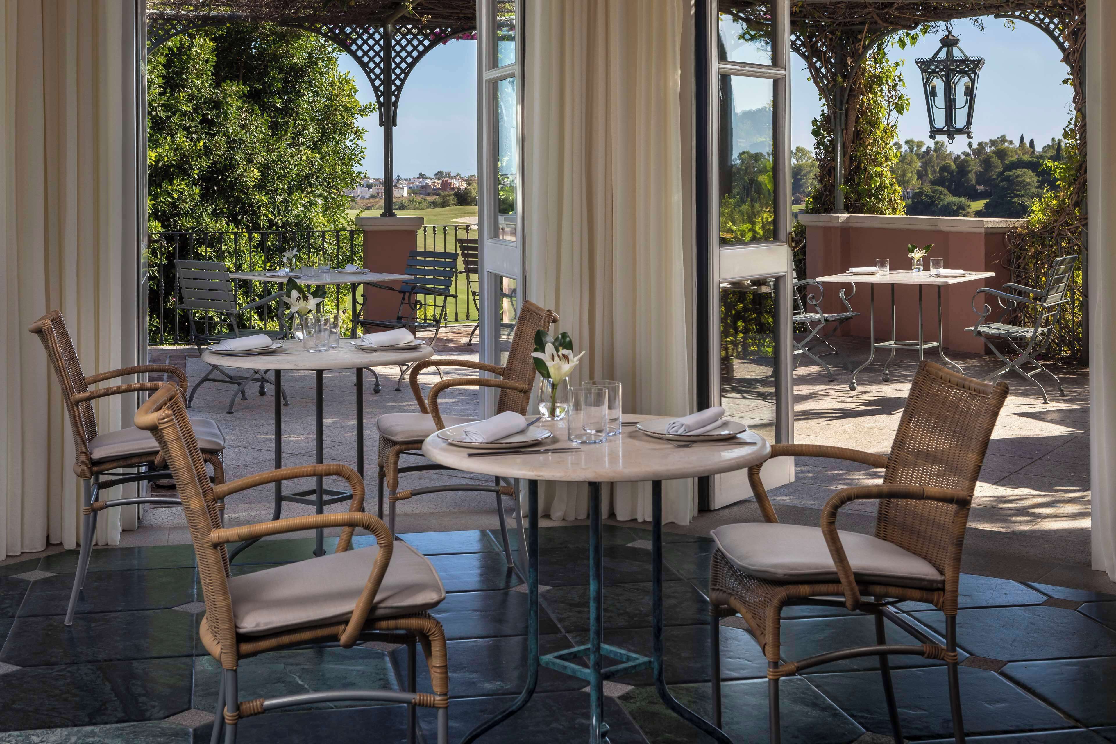 Un área de comedor al aire libre con sillas de mimbre y mesas redondas, con vistas a un jardín exuberante y colinas distantes.