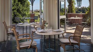 Un área de comedor al aire libre con sillas de mimbre y mesas redondas, con vistas a un jardín exuberante y colinas distantes.