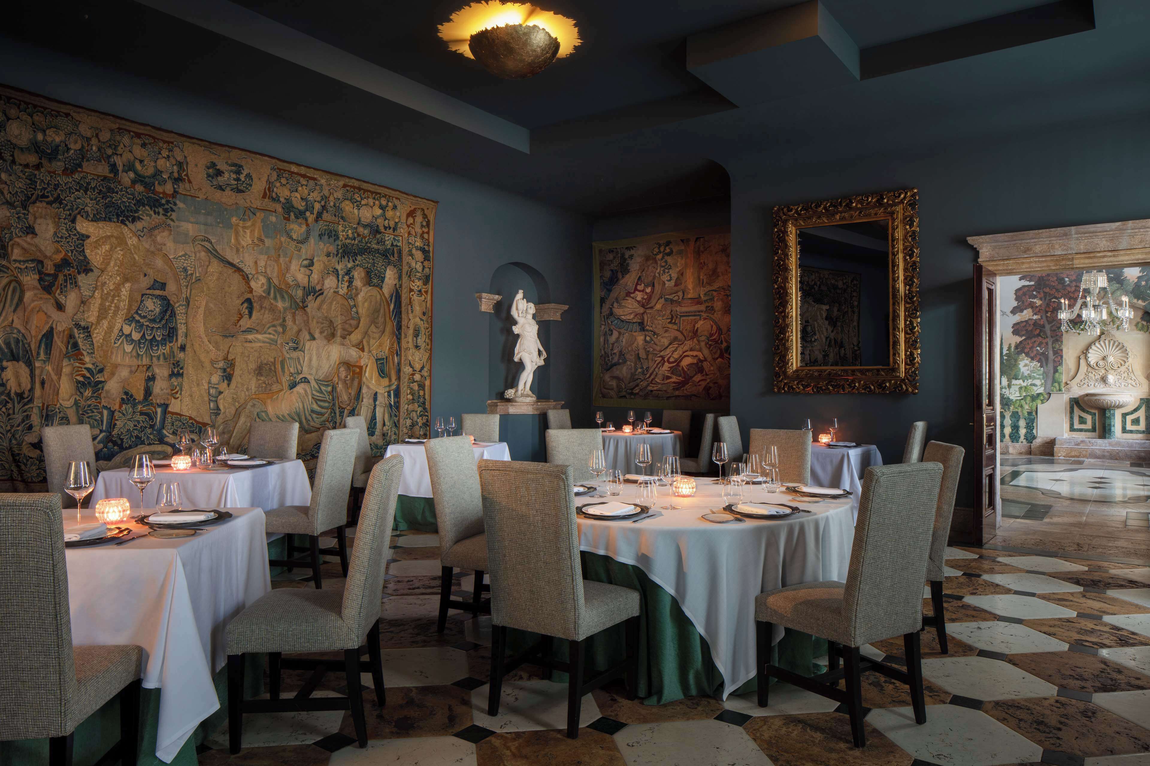 Un comedor con mesas dispuestas para una comida, rodeado de grandes cuadros decorativos y una araña.