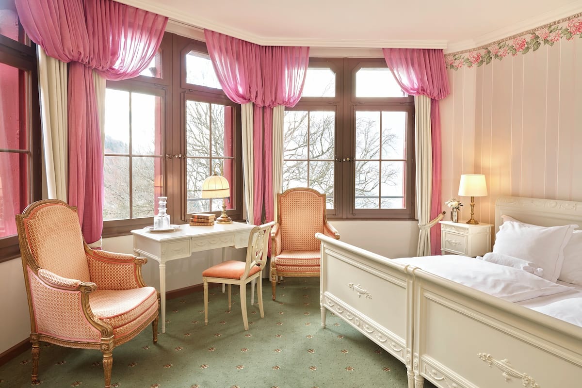 Quarto com cama, duas cadeiras, escrivaninha e janela com cortinas.