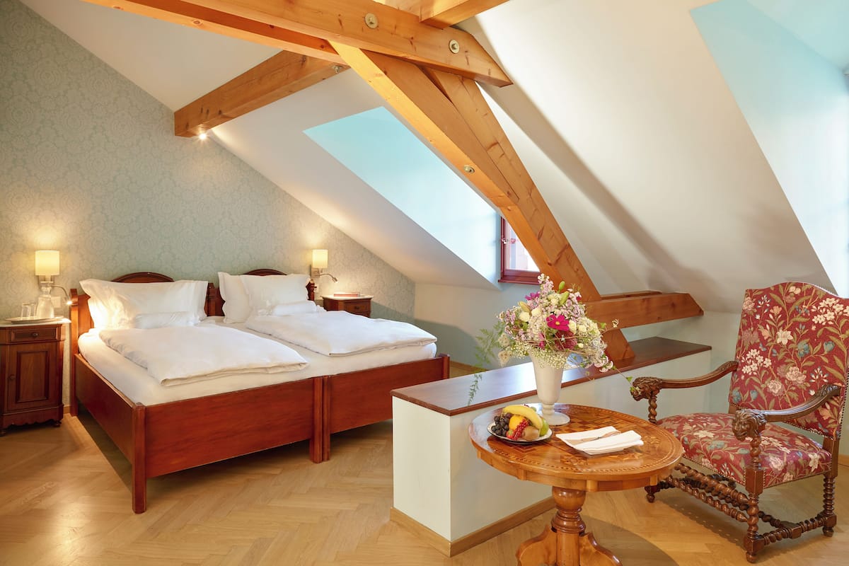 Quarto com cama de madeira, mesa de cabeceira, mesa com frutas, uma cadeira e um arranjo floral.