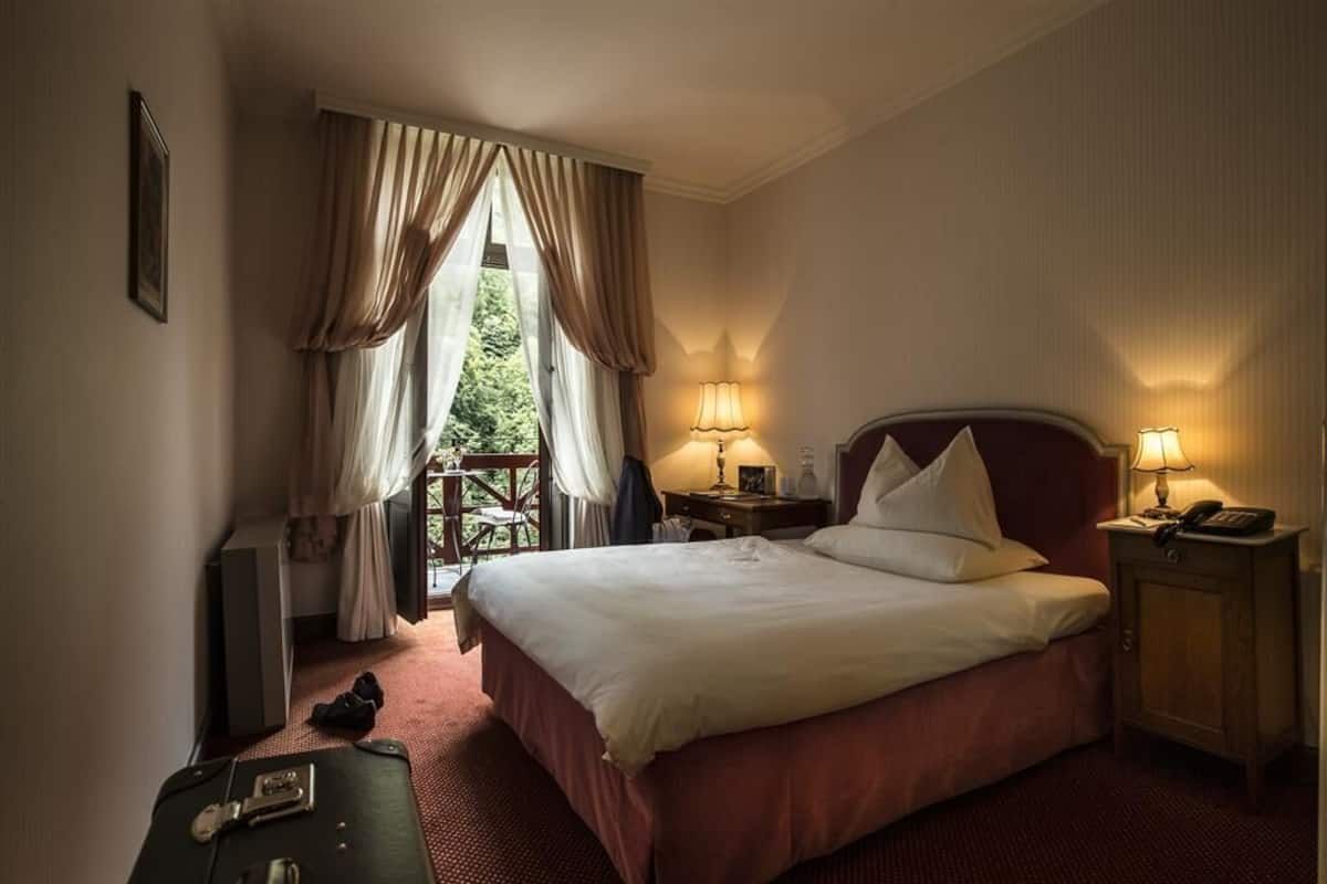 Quarto de hotel com uma cama grande, duas luminárias de cabeceira, uma mesa de cabeceira de madeira, uma escrivaninha com cadeira e vista pela janela.