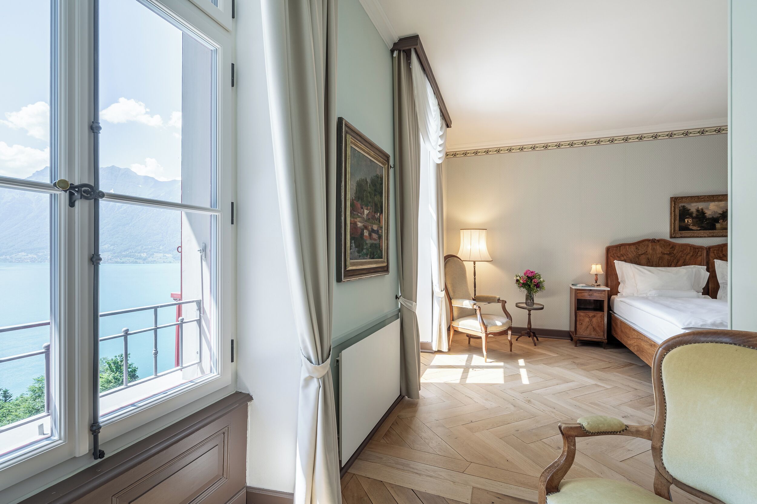 A hotel room with a bed, a chair, a nightstand, a lamp, a painting, and a view of a body of water.