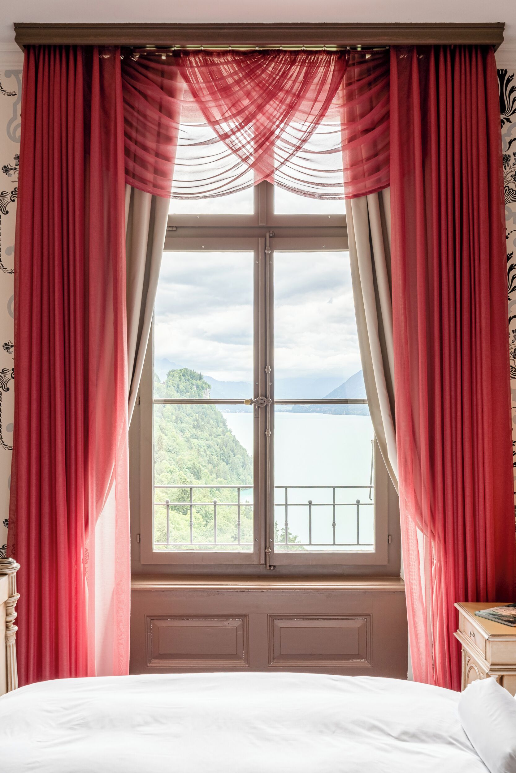 Un dormitorio con un ventanal grande, cortinas rojas y vistas a un paisaje montañoso.