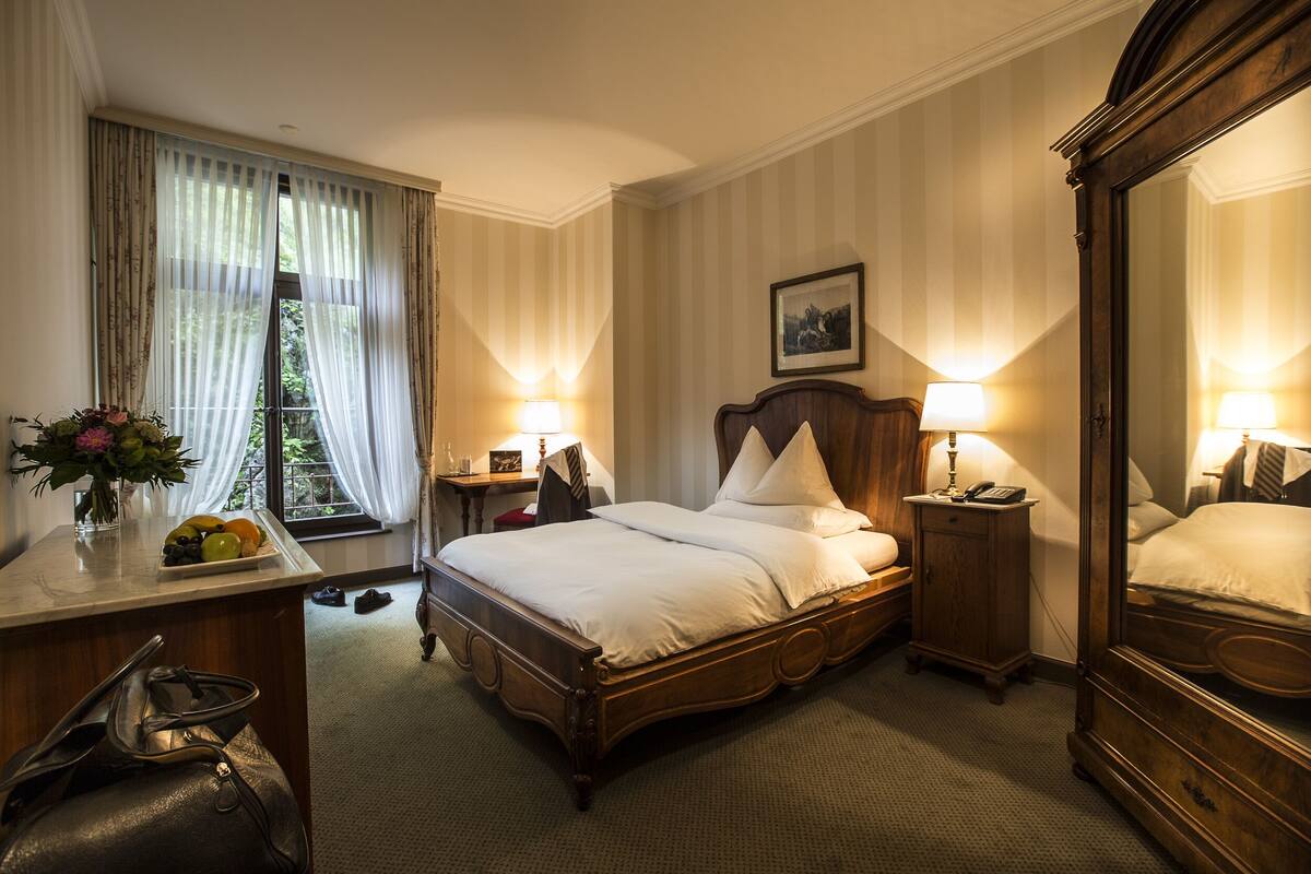 Quarto de hotel com cama de madeira, uma escrivaninha com um vaso de flores, mesa de cabeceira com abajur e um espelho grande.