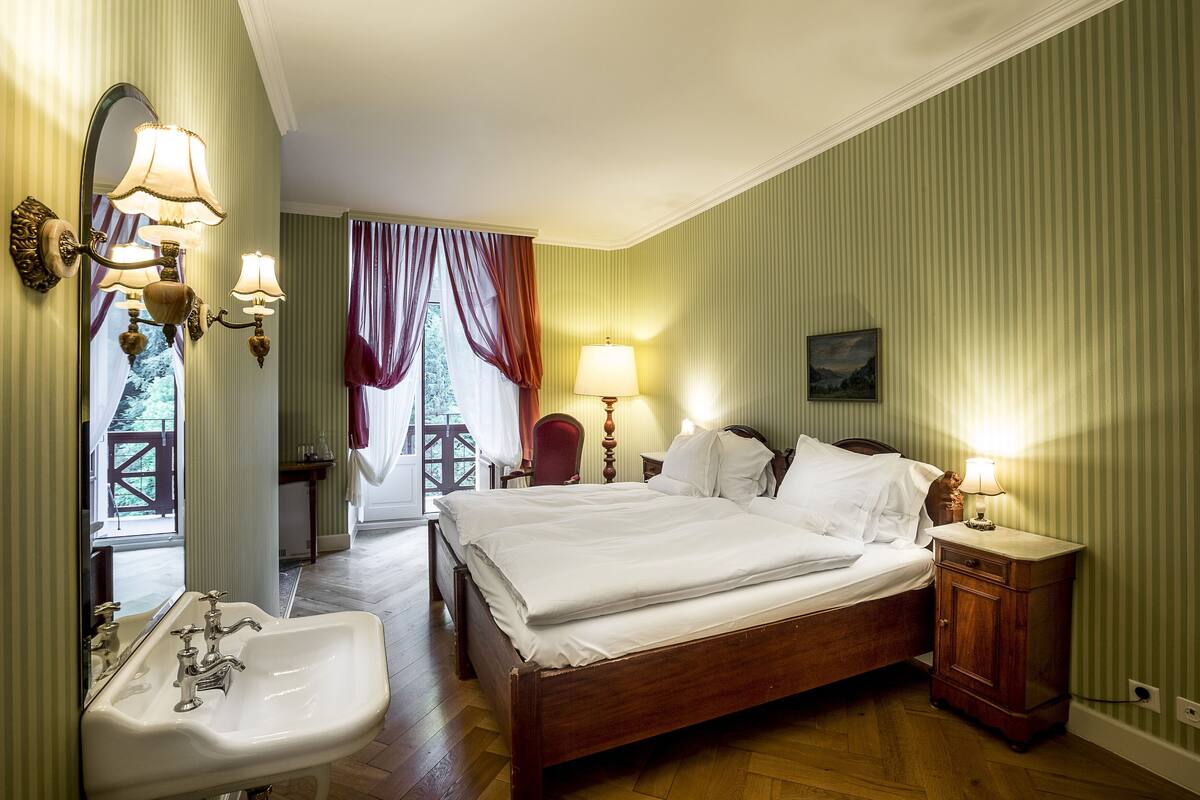 Quarto de hotel com uma cama grande, uma mesa de cabeceira de madeira, um lavatório e uma janela com cortinas.