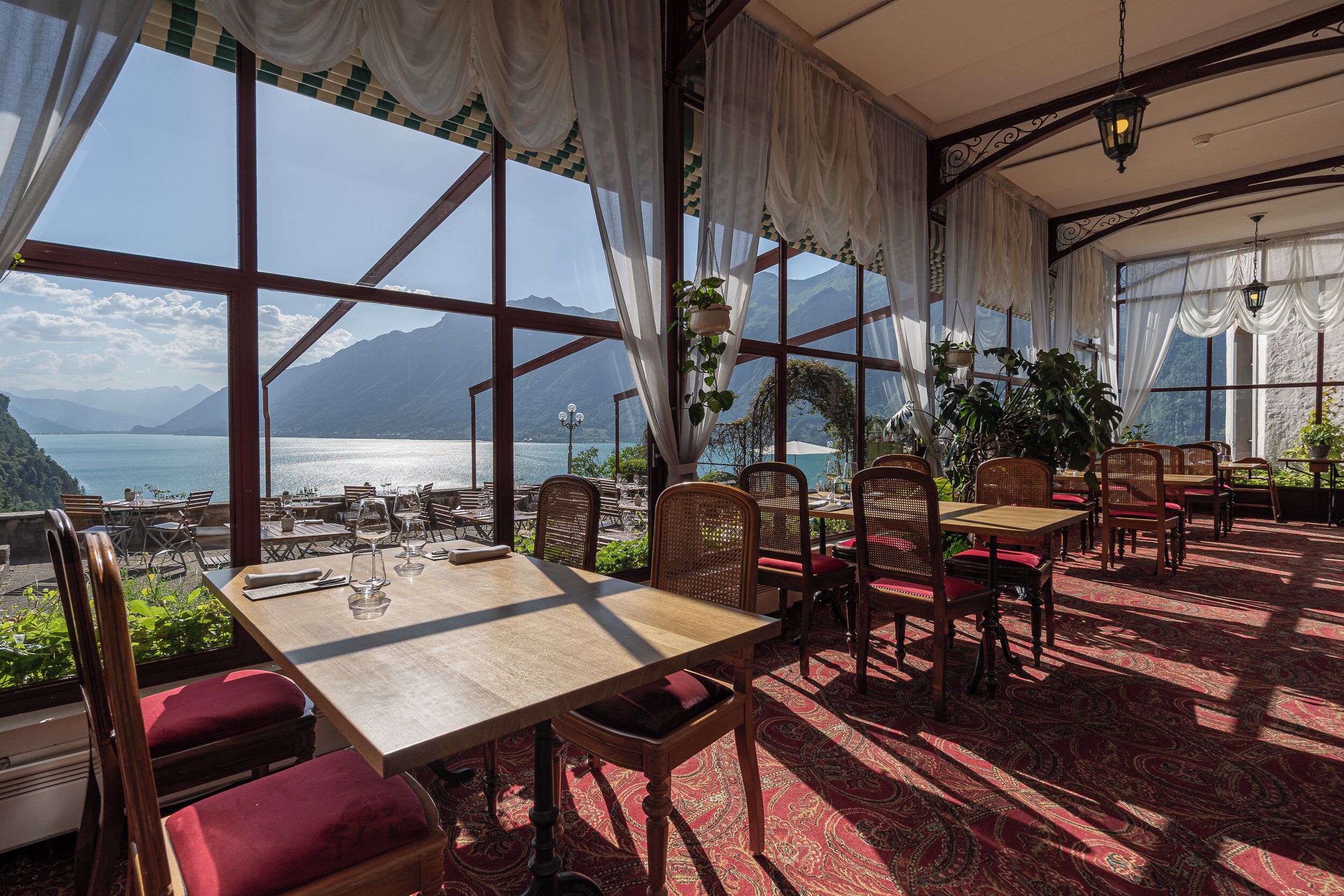 Un restaurante con mesas y sillas dispuestas para comer, grandes ventanales que ofrecen vistas a un lago y montañas, y plantas colgantes.