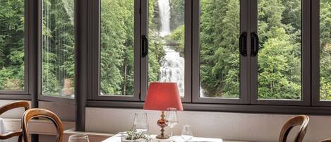A dining table set for two with a view of a waterfall through large windows.