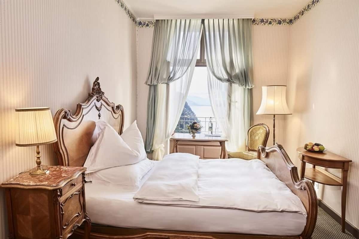 Uma cama clássica com dossel e cabeceira decorada, duas mesas de cabeceira com luminárias, uma mesinha com frutas e vista para o exterior através de cortinas leves.