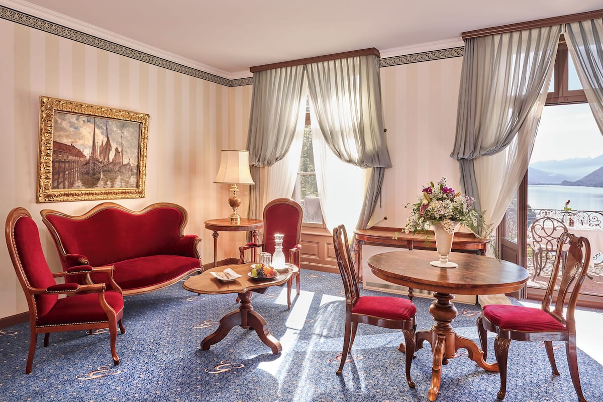 Quarto com sofá vermelho, mesa de centro de madeira e mesa de jantar com cadeiras. Há uma pintura na parede e vista para um corpo d'água pela janela.