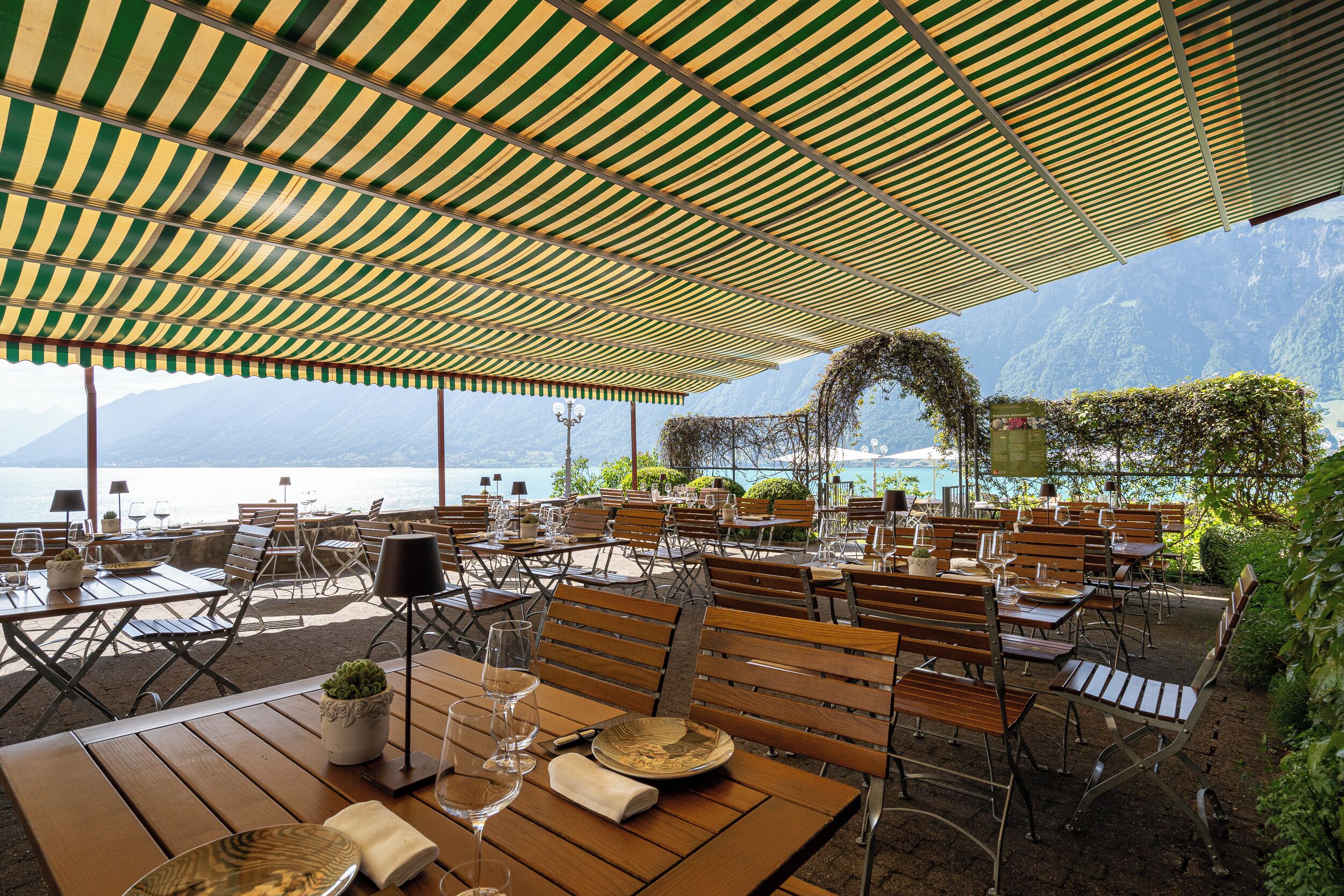 An outdoor dining area with tables and chairs, a canopy overhead, and a view of the sea and mountains in the background.