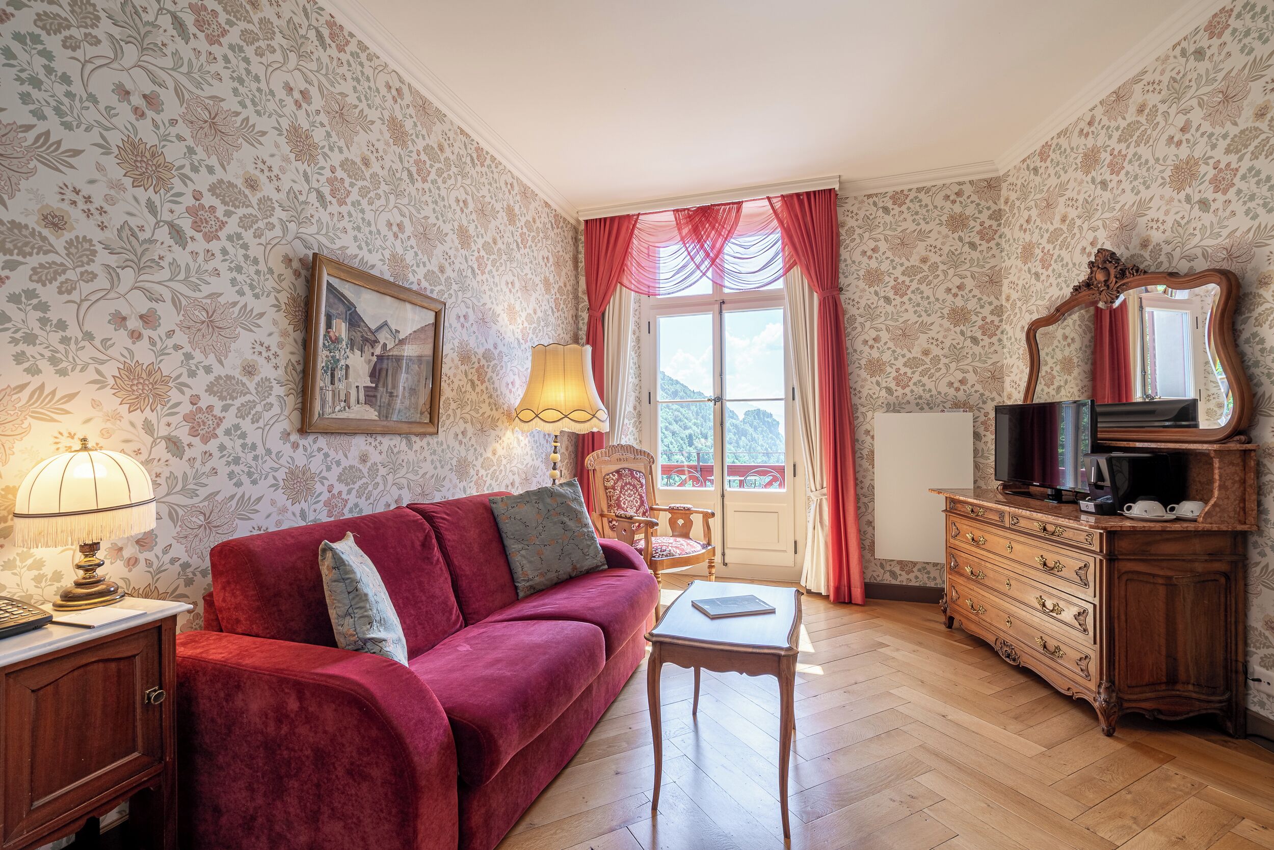 A room with a red sofa, a wooden dresser, a television, a lamp, and a window with curtains.