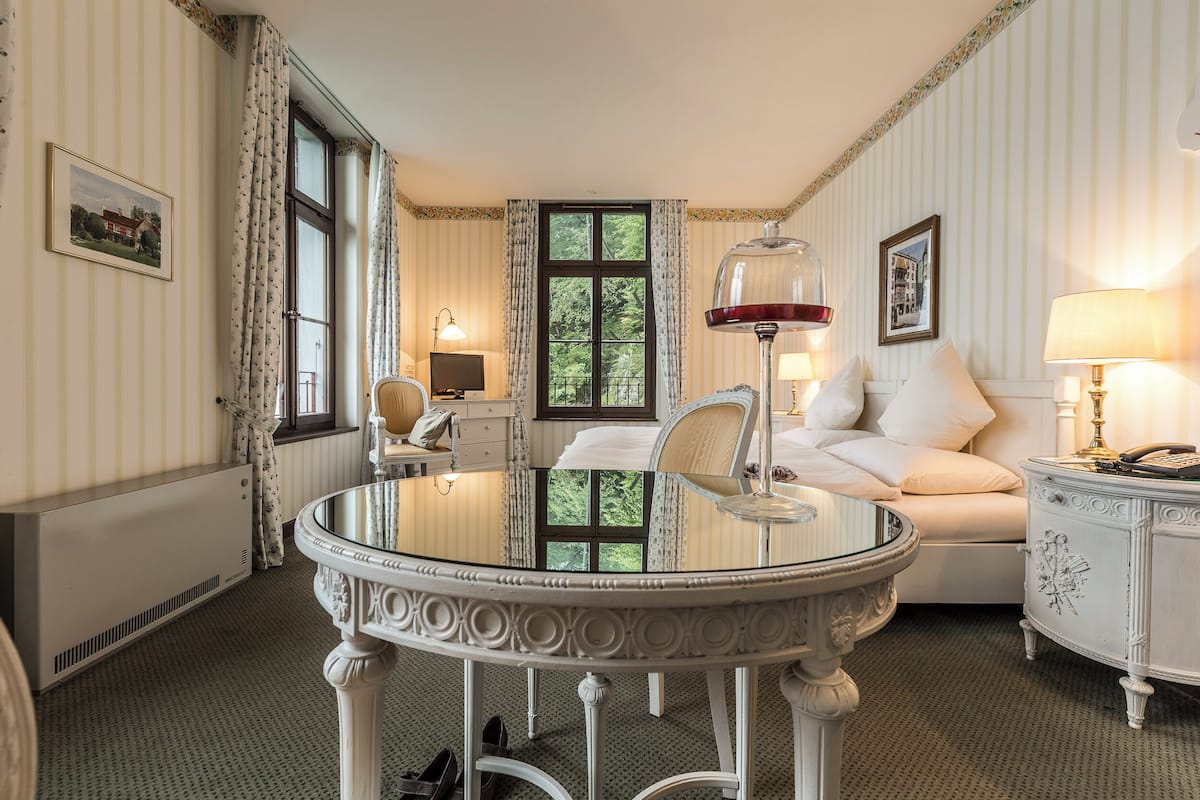 Quarto de hotel com uma mesa redonda, uma cama com roupa branca, uma mesa de cabeceira com abajur e vista para o verde pela janela.