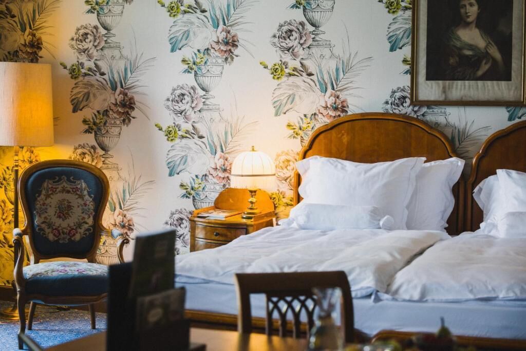 Une chambre d’hôtel avec un grand lit, une table de chevet avec une lampe, une chaise et un papier peint à motifs floraux.