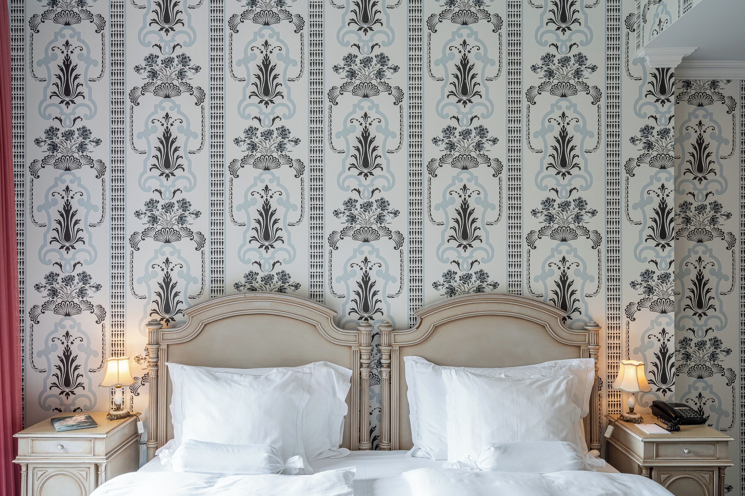 Habitación de hotel con cama clásica, dos mesitas de noche con lámparas y un papel tapiz decorativo con motivos florales.