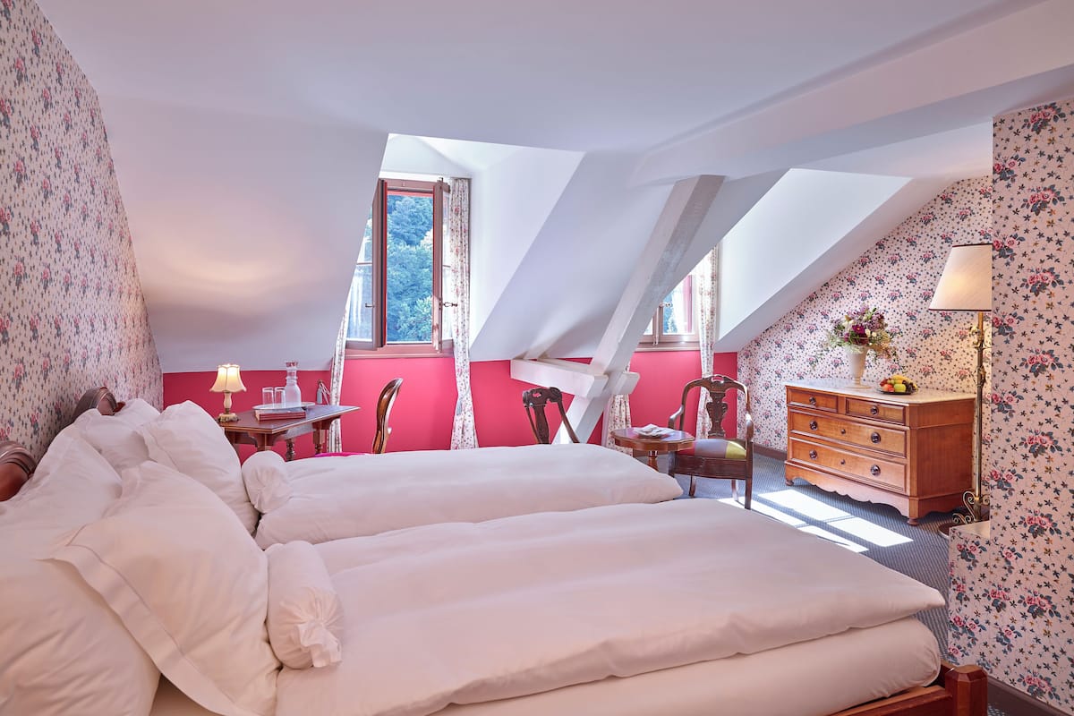 Quarto com duas camas, cômoda, luminária e janela com vista para árvores.
