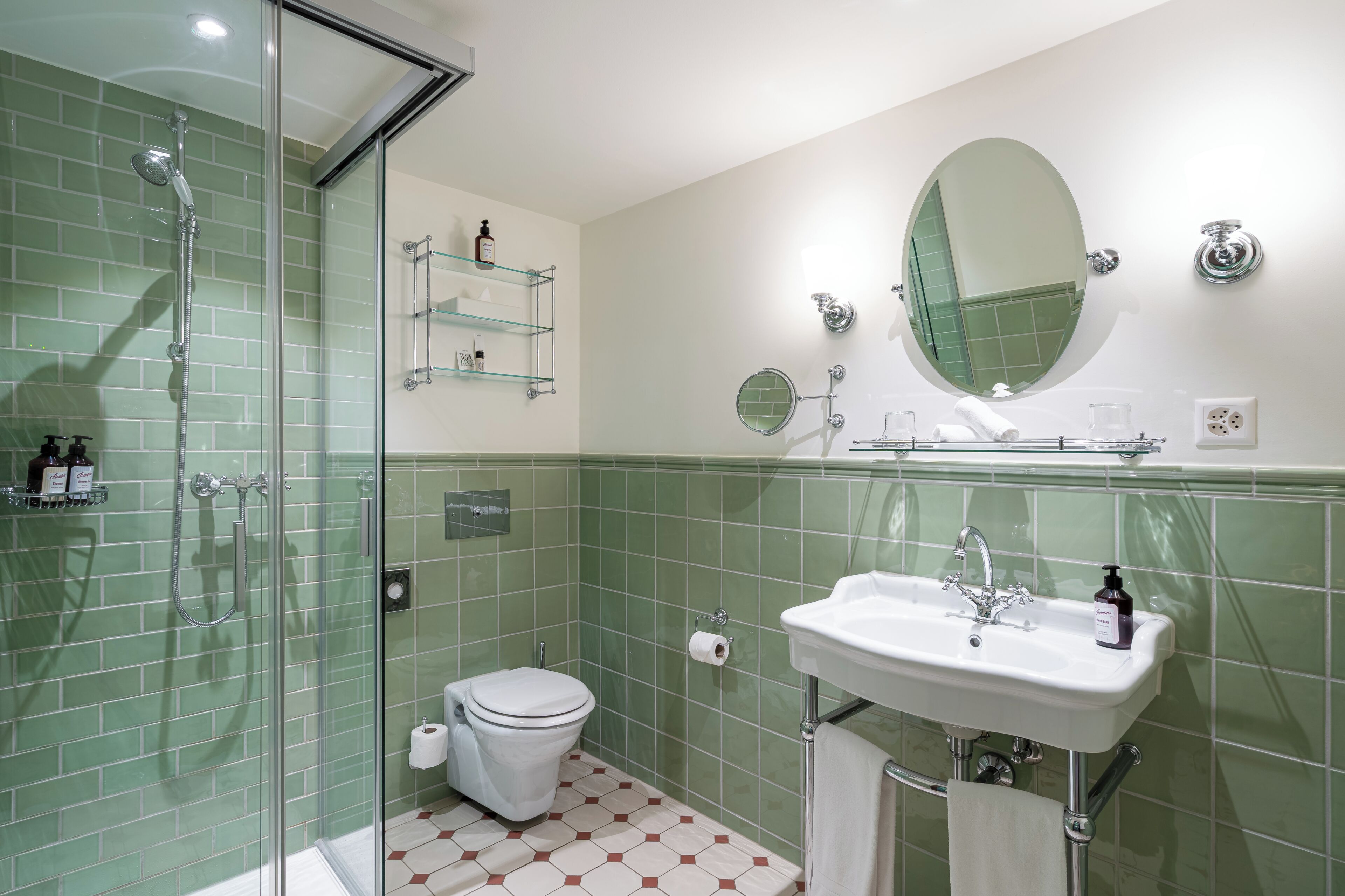 Un baño con cabina de ducha de vidrio, un inodoro blanco, un lavabo con espejo y paredes con azulejos verdes.