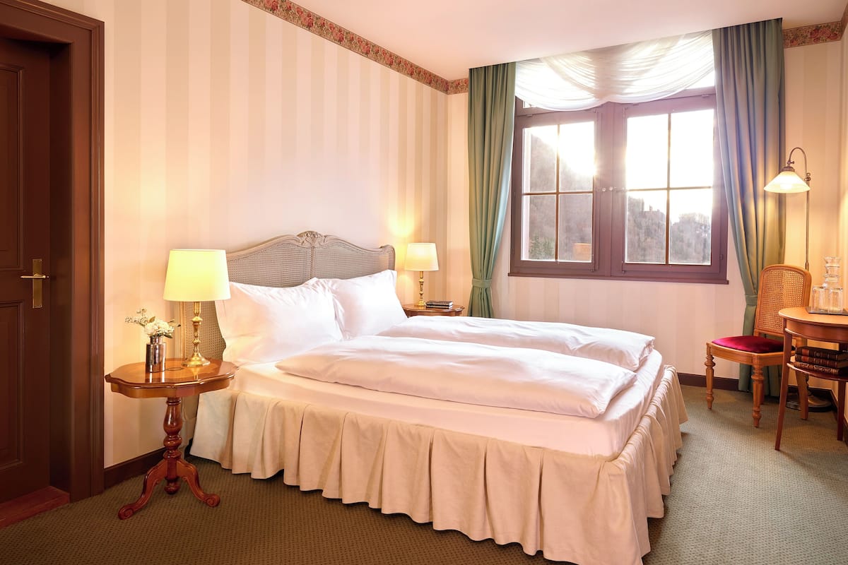 Quarto de hotel com uma cama grande, duas mesas de cabeceira com luminárias, uma cadeira, uma mesinha com livros e uma janela com cortinas.