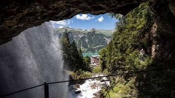 Eingang einer Höhle mit Blick auf einen Wasserfall, eine Berglandschaft und ein Gebäude in der Ferne.