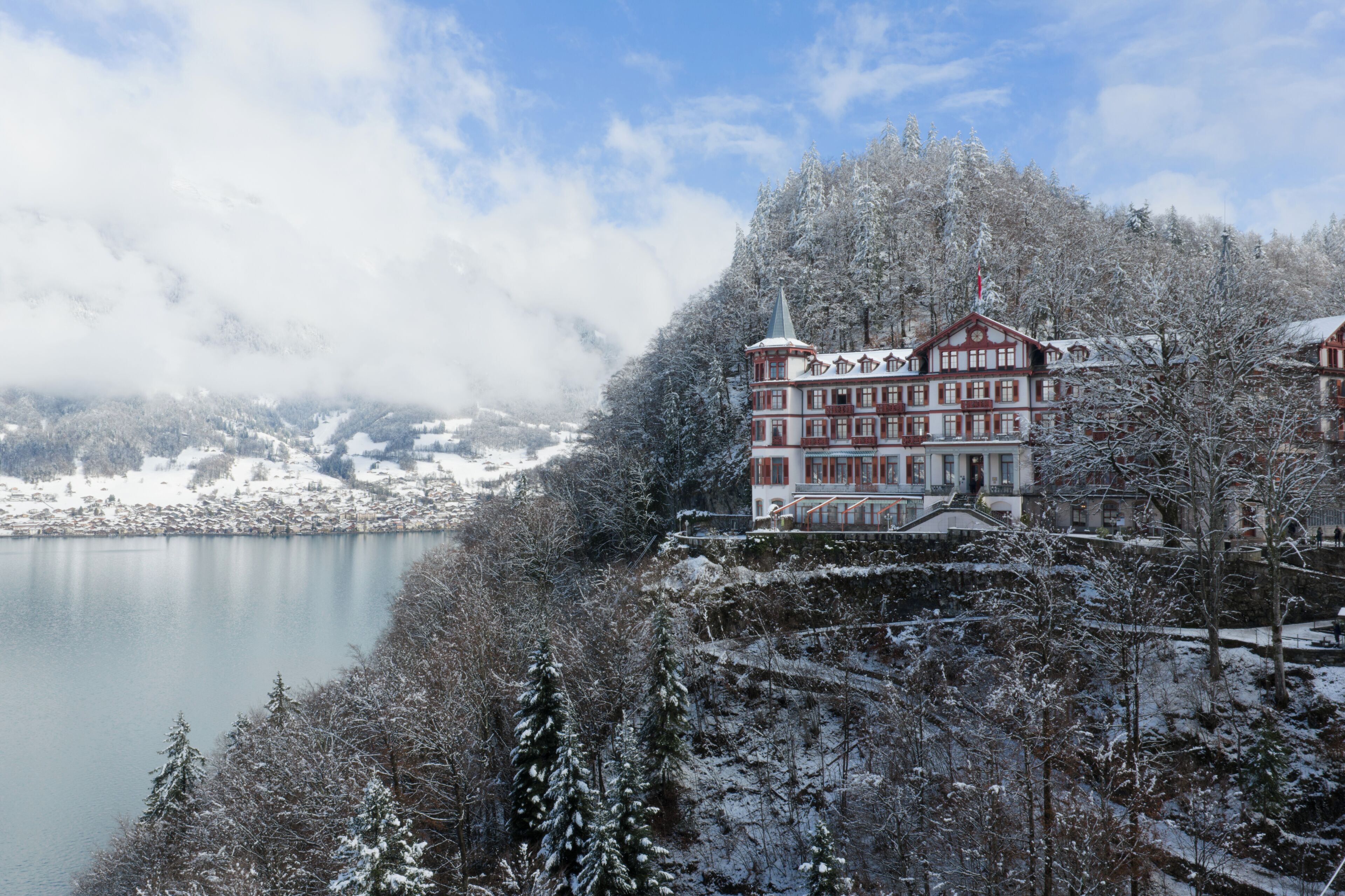 A snow-covered building with a tower, surrounded by a lake and forested mountains.